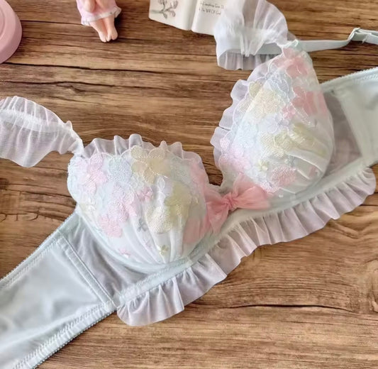 A pastel lace bra with ruffled trim and floral applique in soft pink, white, and pale yellow, laid flat on a wooden surface with matching underwear pieces nearby.