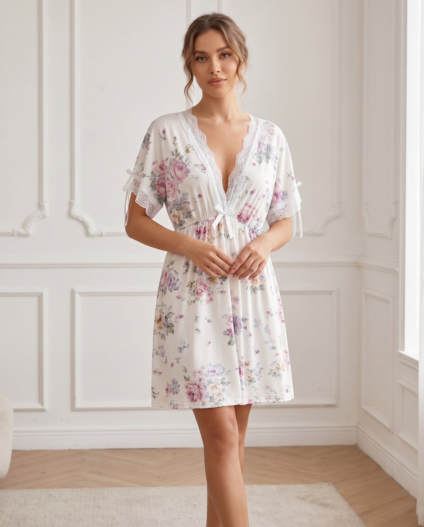 A woman wearing a short white floral nightgown with lace trim and a tie waist, standing in a bright room with white paneled walls.