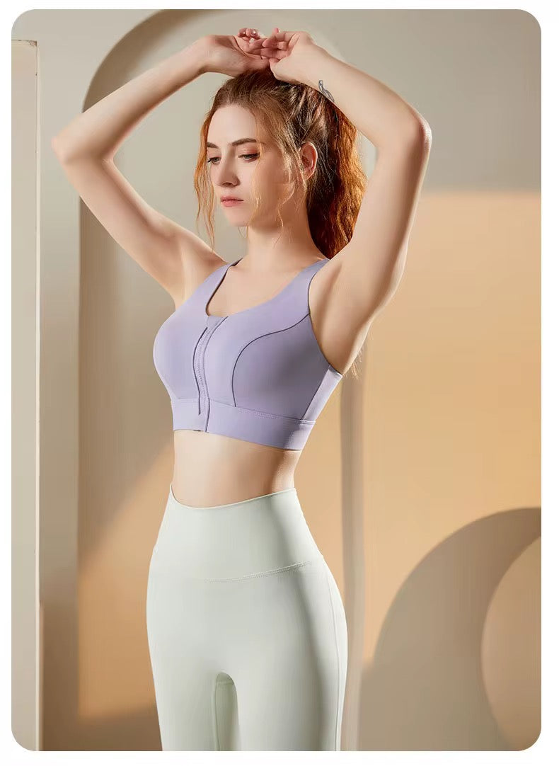 A woman wearing a lavender athletic sports bra and light green leggings poses with her hands behind her head in a warm, minimalist studio setting.