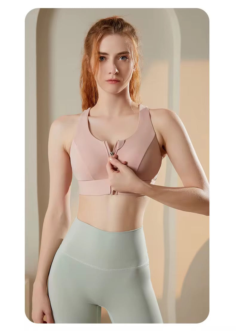 A woman modeling a light pink sports bra with a front zipper and matching underband, paired with high-waisted light sage green leggings, set against a warm beige background with soft arches.