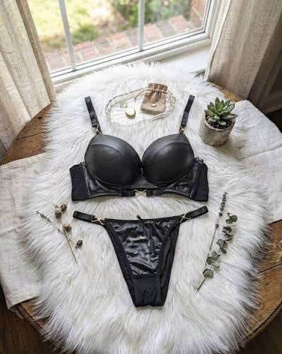 A black satin lingerie set consisting of a bra and matching panties, styled in a flat-lay arrangement on a white faux fur rug next to a window.
