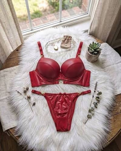 A shiny red two-piece lingerie set consisting of a bra and matching panties, styled on a white faux fur rug on a wooden table with a window in the background.