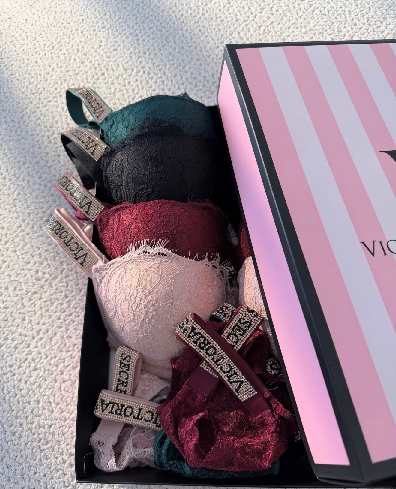 A Victoria's Secret gift box containing multiple lace bralettes in black, teal, blush pink, and burgundy, next to its pink and white striped box on a textured white fabric background.