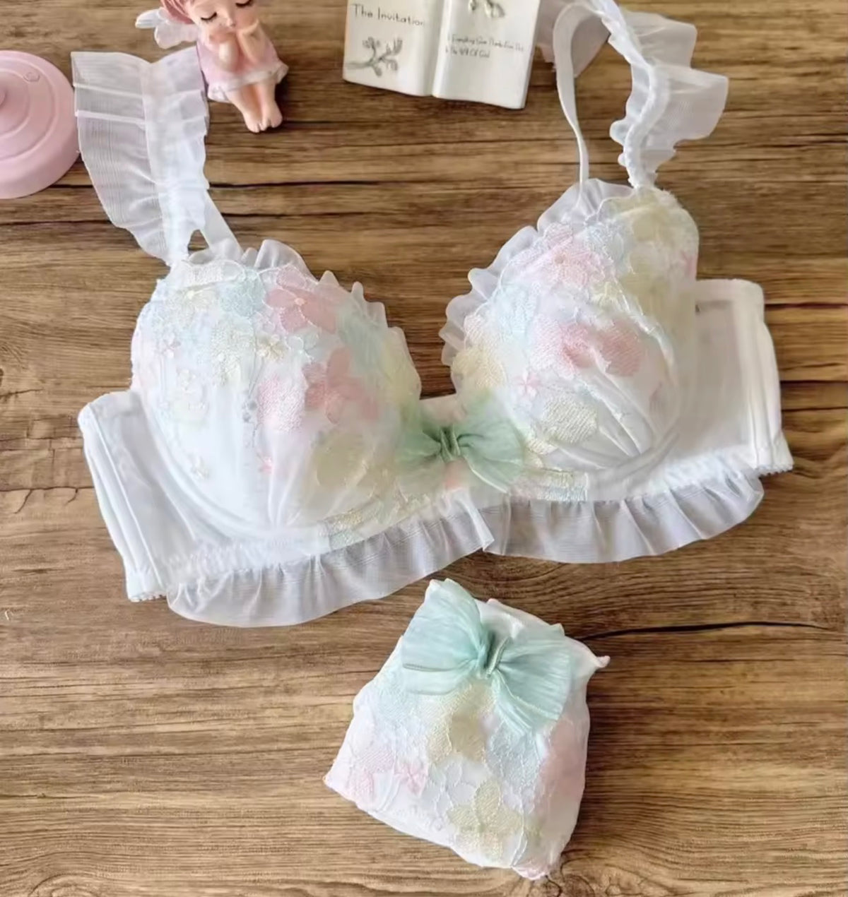 A pastel-colored lace bra and matching underwear set arranged on a wooden surface with decorative props, including a small doll, a book, and a pink cup.