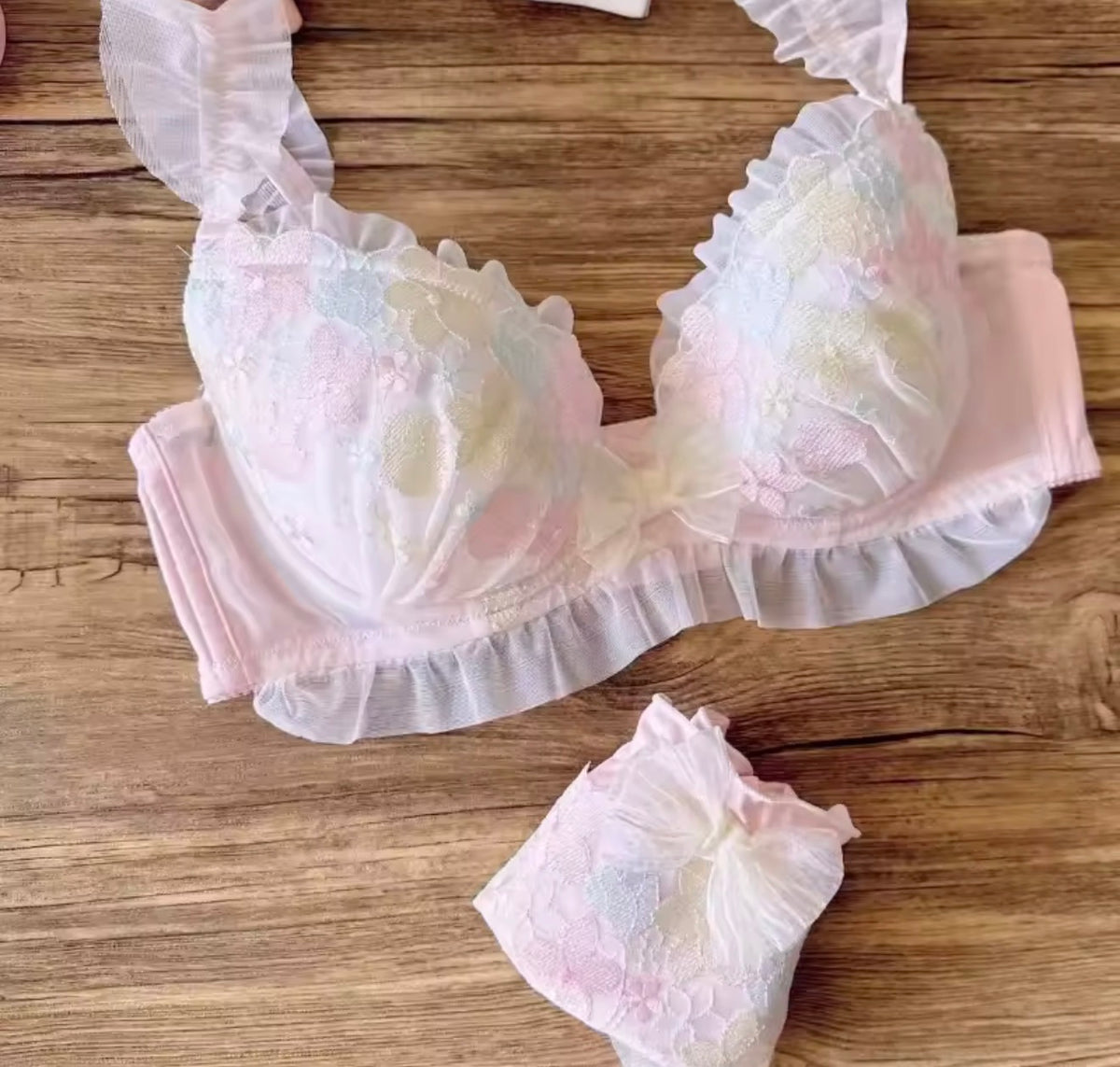 A pastel pink lace baby bra with ruffled trim and a matching folded diaper, displayed on a wooden surface.