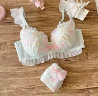 A pastel pink and white ruffled lingerie set laid flat on a wooden surface, including a bralette with matching panties and a small pouch or box.