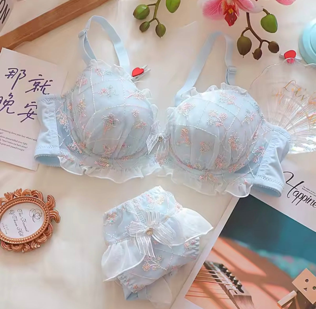 A light blue lace bra and matching panties set with floral embroidery, displayed in a styled flat-lay arrangement with decorative props including a fan, card, and flowers.