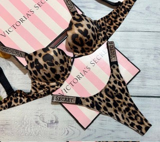 A Victoria's Secret leopard print bralette and matching thong laid flat on pink striped branded boxes over a light wood surface.