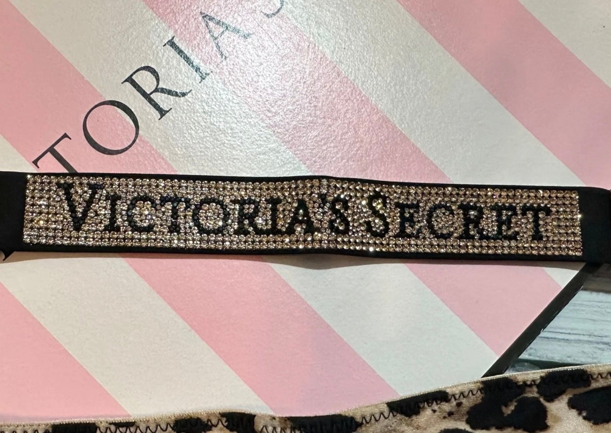 A Victoria's Secret rhinestone-embellished strap or band laid on a pink and white striped box. The strap features black lettering spelling 'VICTORIA'S SECRET' set in a dense row of clear rhinestones on a black base.