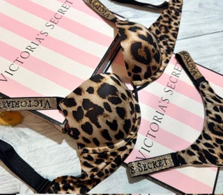A leopard print Victoria's Secret bra laid flat on pink striped Victoria's Secret packaging boxes.
