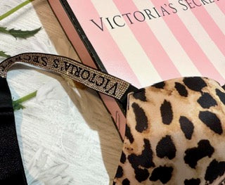 A close-up of a leopard print bra strap with 'VICTORIA'S SECRET' text, resting on a pink and white striped Victoria's Secret box.
